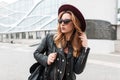 Sexy modern hipster woman in a luxurious purple hat in a fashionable black jacket with a backpack in stylish sunglasses walks Royalty Free Stock Photo