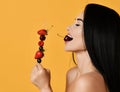 Sexy naked brunette woman holds a skewer with berries and eats bites cherry enjoys nature taste isolated on yellow. Royalty Free Stock Photo