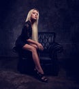 Sexy slim blod model sitting in fashion armchair in black dress and posing on dark dramatic background