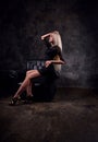 Sexy slim blod model sitting in fashion armchair in black dress and posing on dark dramatic background