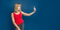 Sexy slim blonde womanin red t-shirt on a blue studio background making selfie with smartphone. Copyspace