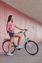 stylish brunette girl wearing a pink flannel shirt and denim shorts in glasses, posing with city bike against a Royalty Free Stock Photo