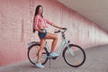 stylish brunette girl wearing a pink flannel shirt and denim shorts in glasses, posing with city bike against a Royalty Free Stock Photo