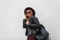 Sexy urban hipster young woman in a fashionable hat in stylish sunglasses in a vintage leather jacket with a backpack walks Royalty Free Stock Photo
