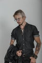 Sexy young blond man with tattoos in black trendy denim clothes with a jacket in round glasses posing in the studio near a vintage Royalty Free Stock Photo