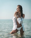 Sexy young slim woman girl in white bikini and shirt stands in sea touching her shoulder feeling softness. Summer vibes Royalty Free Stock Photo