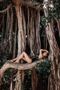 Sexy young woman with big breast in black swimsuit is lying on big branch of tree in tropical forest. Summer vibes