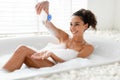 Sexy young woman lying in bubble bath near window, relaxing, adding foam into water, taking care of her body at home