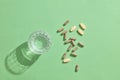 A shadow-casting glass of water placed next to scattered pastel-colored pills on the floor against a green background.