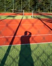 Shadow of a man making photo on a tennis court. Red polyurethane surface of the sports court. Trees in the background. Sunny day