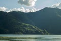 A shadow side of the mountain with green forest and houses under the blue sky with white clouds on the background of the lake Royalty Free Stock Photo