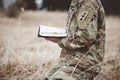 Shallow focus shot of a young soldier kneeling while holding an open bible in a field Royalty Free Stock Photo