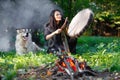 Shaman woman with an Alaskan Malamute drumming in the evening forest by the fire