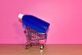 Shampoo in shopping trolley on table on pink background. Blue liquid in a plastic bottle in a basket cart. Detergent bottle for