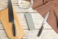 Sharpening stone, knives and water on white wooden table, flat lay