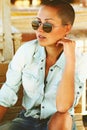 Shaved-headed girl facial portrait, suntan face, black stylish sunglasses, fashion jeans clothes t-shirt and shorts, brown skin. B