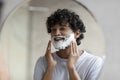 Shaving routine concept. Indian man applying shave foam on face making morning hygiene and looking at his reflection Royalty Free Stock Photo
