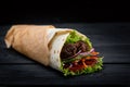 Shawarma rolled in lavash, moist grilled meat with onion, herbs and vegetables on wooden black background.