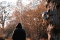 A sheep skull hanging from a tree, while a sinister hooded figure stands in the background, blurred and out of focus. With a