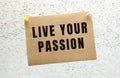 A sheet of craft paper with LIVE YOUR PASSION text attached to a white textured wall with a button. Office reminder. Royalty Free Stock Photo