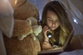 Shes a dedicated bookworm. a young girl reading a book by torchlight under a blanket fort at home.