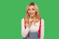 Shh, don`t speak! Portrait of positive adult blond woman in stylish overalls holding finger on lips in silence gesture