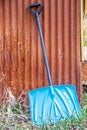 Shinny blue metal show shovel leaning against a grainy rusty corregated shed with green and dry grasses growing at the bottom