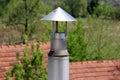 Shiny metal narrow chimney with damaged homemade protective cover on top with houses and dense trees in back