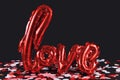 Shiny red balloon forming the word `love` on dark black background with colorful confetti on ground