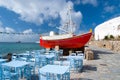 Ship and tavern furniture on quay in Mykonos, Greece. Red boat and blue tables on sea beach. Beach restaurant with sea