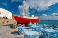 Ship and tavern furniture on quay in Mykonos, Greece. Red boat and blue tables on sea beach. Beach restaurant with sea