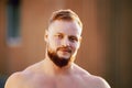 Shirtless broad shouldered bearded white man in his 30s outdoors close up.