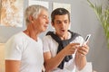 Shocked adult man sitting with his senior father in living room at home using smartphone see shock content browsing internet web Royalty Free Stock Photo