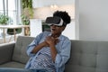 Shocked African American woman in virtual reality glasses open mouth sits on sofa in living room Royalty Free Stock Photo