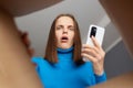Shocked amazed Caucasian woman unpacking box and looking in cardboard box with open mouth and big eyes holding mobile phone in Royalty Free Stock Photo