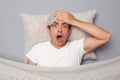 Shocked amazed man with resting on white pillow under blanket wearing t-shirt isolated over grey background looking nat camera Royalty Free Stock Photo