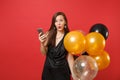 Shocked amazed young girl in little black dress holding air balloons using mobile phone while celebrating isolated on Royalty Free Stock Photo