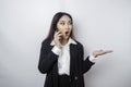 Shocked Asian businesswoman wearing a black suit pointing at the copy space beside her while talking on her phone, isolated by a Royalty Free Stock Photo