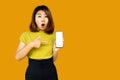Shocked Asian woman hand pointing at smart phone with surprise face standing over orange background
