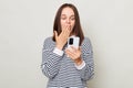 Shocked astonished brown haired adult woman wearing striped shirt standing isolated over gray background using smart phone