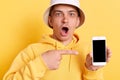 Shocked astonished man wearing casual style hoodie and panama posing isolated over yellow background, showing smart phone with Royalty Free Stock Photo