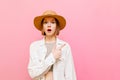 Shocked attractive girl stands against a pink background in a hat and white summer clothes,looks in camera and points finger to