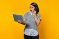 Shocked beautiful young woman talking on smartphone and looking at laptop screen on yellow background Royalty Free Stock Photo