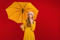 Shocked beautiful young woman with an umbrella in a warm hat on her head, on a red background Royalty Free Stock Photo