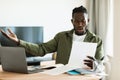 Shocked black man reading paper letter mail and spreading hands, sitting at table with laptop at home office Royalty Free Stock Photo