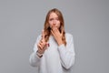 Shocked blonde woman pointing finger up, covers her mouth hand, oh no you did not do, wears white sweater, gray background Royalty Free Stock Photo
