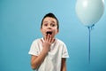 Shocked boy covers his mouth while looking at camera and expressing surprise standing on blue background with copy space