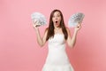 Shocked bride woman with opened mouth in white wedding dress holding bundle lots of dollars, cash money on pink