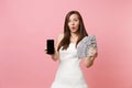 Shocked bride woman in wedding dress holding bundle lots of dollars cash money, mobile phone with blank black empty