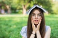 Shocked woman sitting in park with book on head while holding her arms on cheeks and looking at the camera Royalty Free Stock Photo